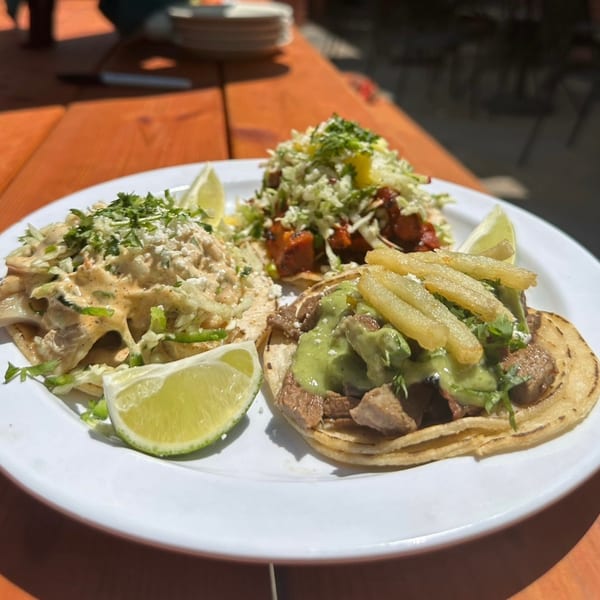 Three vibrant Uno Mas tacos on a white plate, garnished with lime wedges, on an orange table.