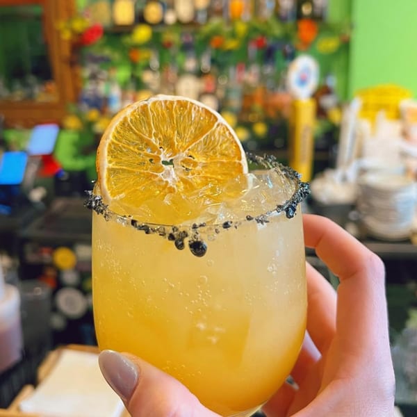 Hand holding a vibrant yellow Uno Mas Taqueria y Cantina cocktail with a dried orange slice and dark rim.