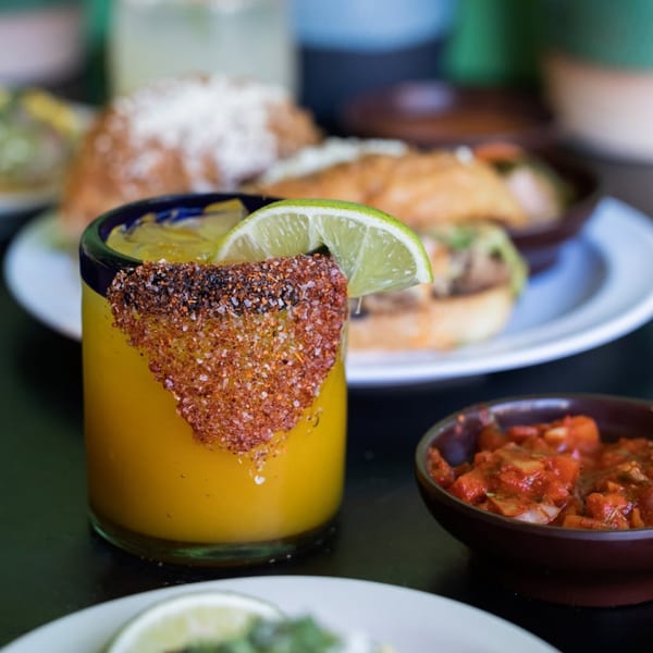A vibrant yellow drink with a chili-salt rim and lime wedge, next to a bowl of salsa.