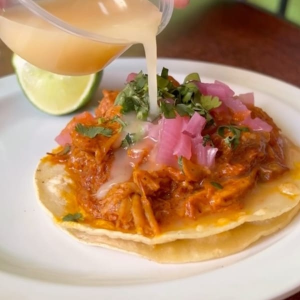 A hand pours a light sauce over a chicken taco with pickled onions, cilantro, and a lime wedge.