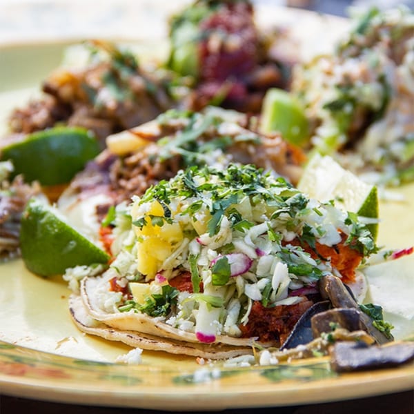 Close-up of multiple Uno Mas Taqueria y Cantina tacos with various toppings and lime wedges.