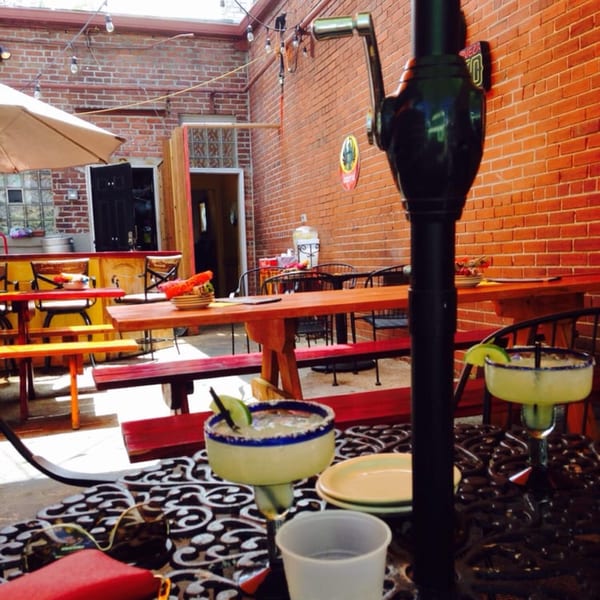 Outdoor patio with brick walls, wooden tables, red benches, and two margaritas on a wrought iron table.