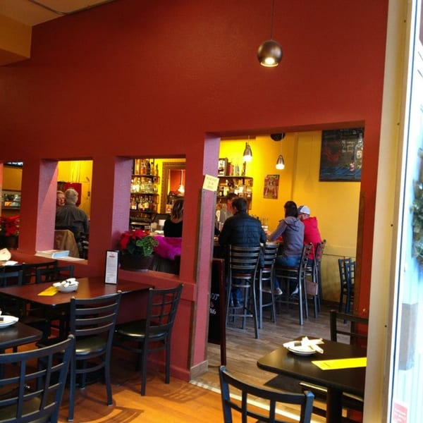A restaurant interior with red walls, black tables, and patrons seated at a bar and dining tables.
