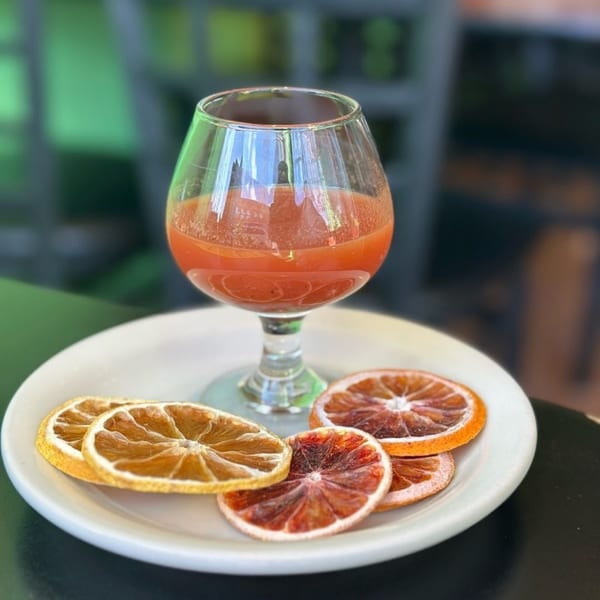 A vibrant red drink in a stemmed glass, served with dried orange and blood orange slices on a white plate.